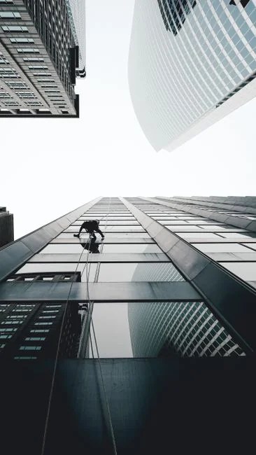 why-choose-us A man cleaning skyscraper windows with a harness, showcasing urban high-rise work.